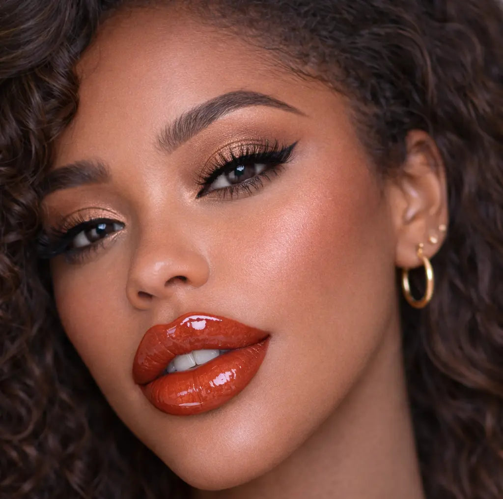 Close-up of a woman with curly hair wearing diamond glow crimson cloth lip gloss with gold hoop earrings.