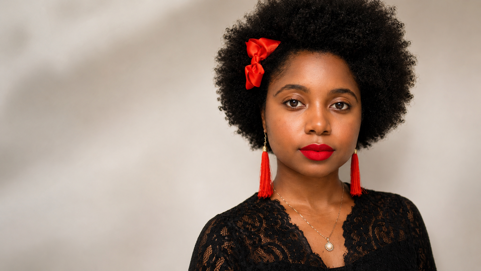 Diamond Michelle with an afro wearing a red ribbon, red earrings, and a red matte lip stain against a neutral background