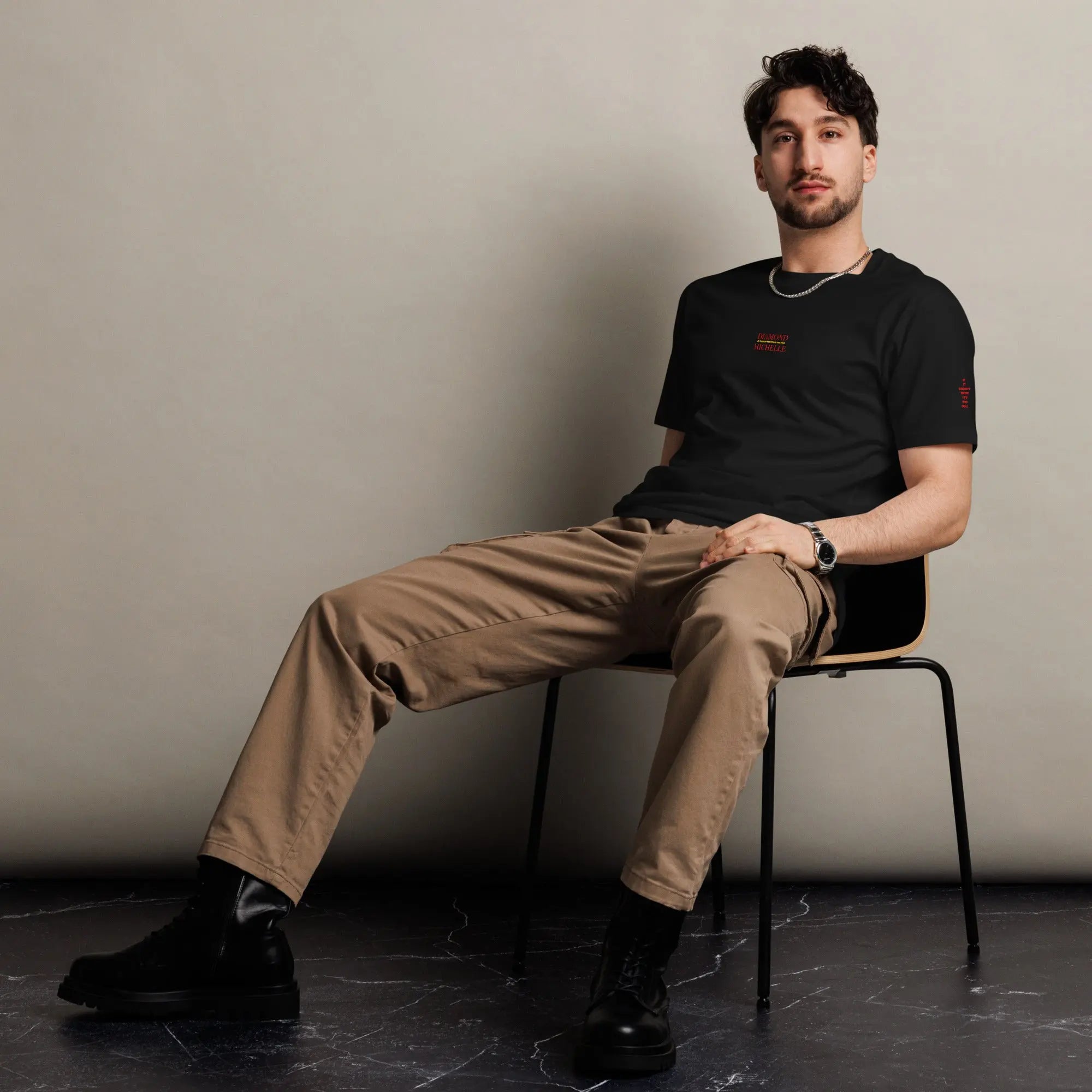 Man sitting on a chair wearing a black t-shirt and beige pants against a plain background